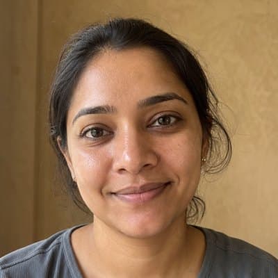 Woman in her thirties with her hair tied back and a soft smile, in a grey-blue T-shirt by a window